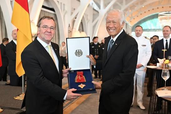 Boris Pistorius presenting an open case containing the insignia of Germany's Order of Merit to a smiling senior guest at a reception in Singapore