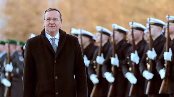 Boris Pistorius in a dark overcoat walking past a line of naval honour guard soldiers in white caps holding rifles