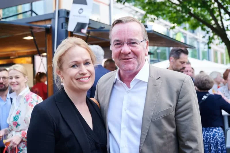 Boris Pistorius smiling in a beige jacket and white shirt at an outdoor summer reception, standing beside a woman in a black blazer
