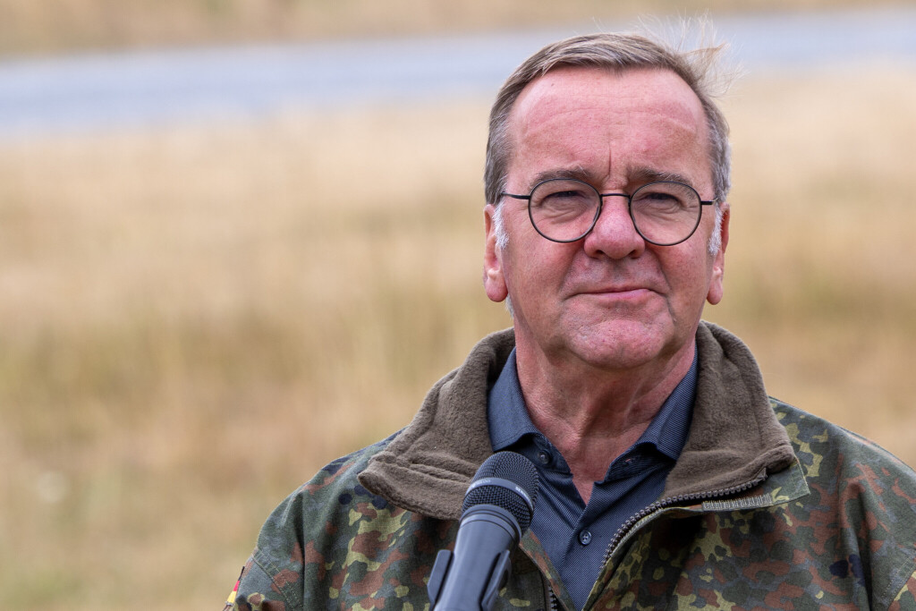 Boris Pistorius in a camouflage field jacket and round glasses standing before a microphone on an open training ground