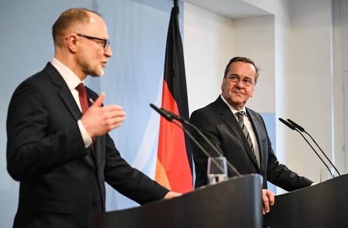 Boris Pistorius standing at a lectern listening as a European counterpart speaks at a joint press conference, with the German flag behind them