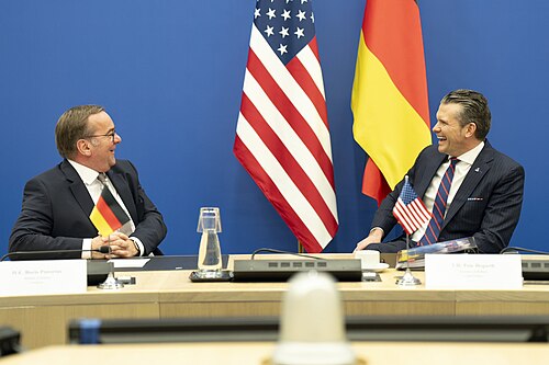 Boris Pistorius and US Defense Secretary Pete Hegseth laughing across a conference table with US and German flags behind them, February 2025
