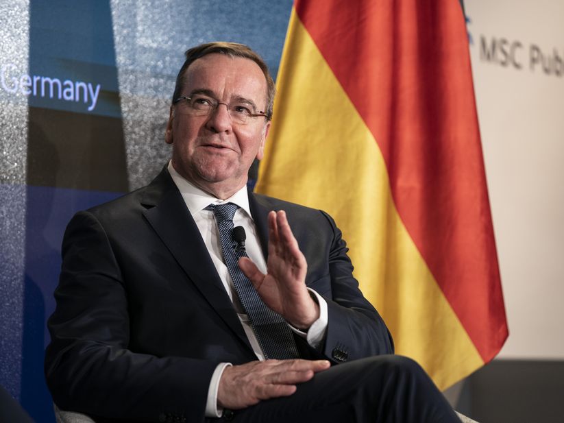 Boris Pistorius seated on stage at a Munich Security Conference event, gesturing with one hand, with a German flag behind him